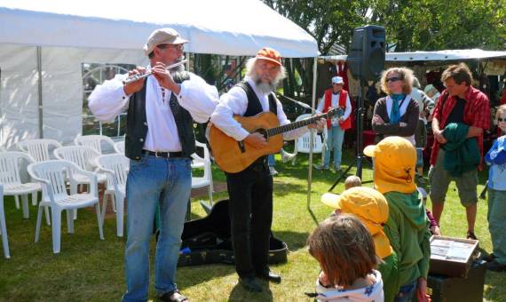 Zwei Musiker mit Flöte und Gitarre spielen auf einem Sommerfest vor Kindern und Erwachsenen unter freiem Himmel